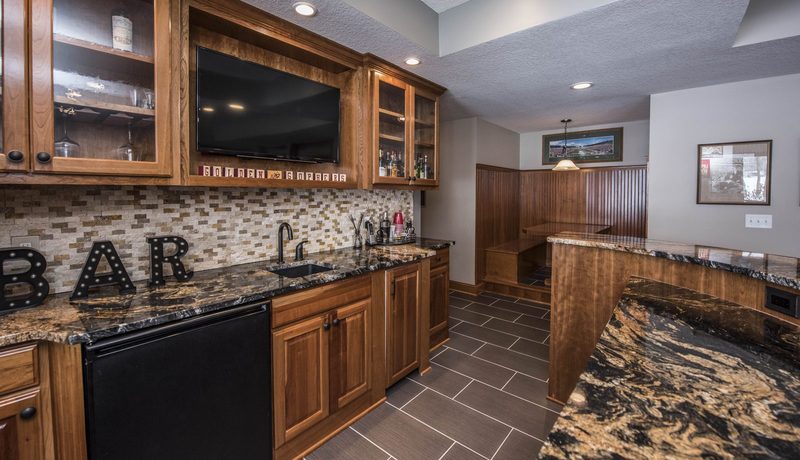 Basement wet bar with cherry cabinetry, stone-tile backsplash, granite counters and a wood-paneled booth